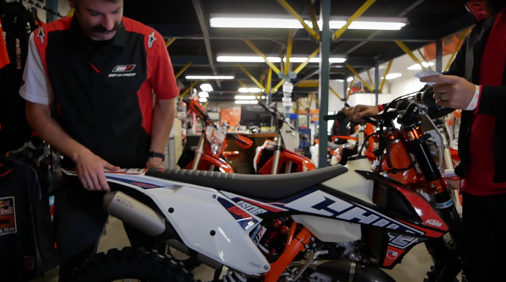 two suspension mechanics test the rebound of a rear shock in an orange KTM dirt bike